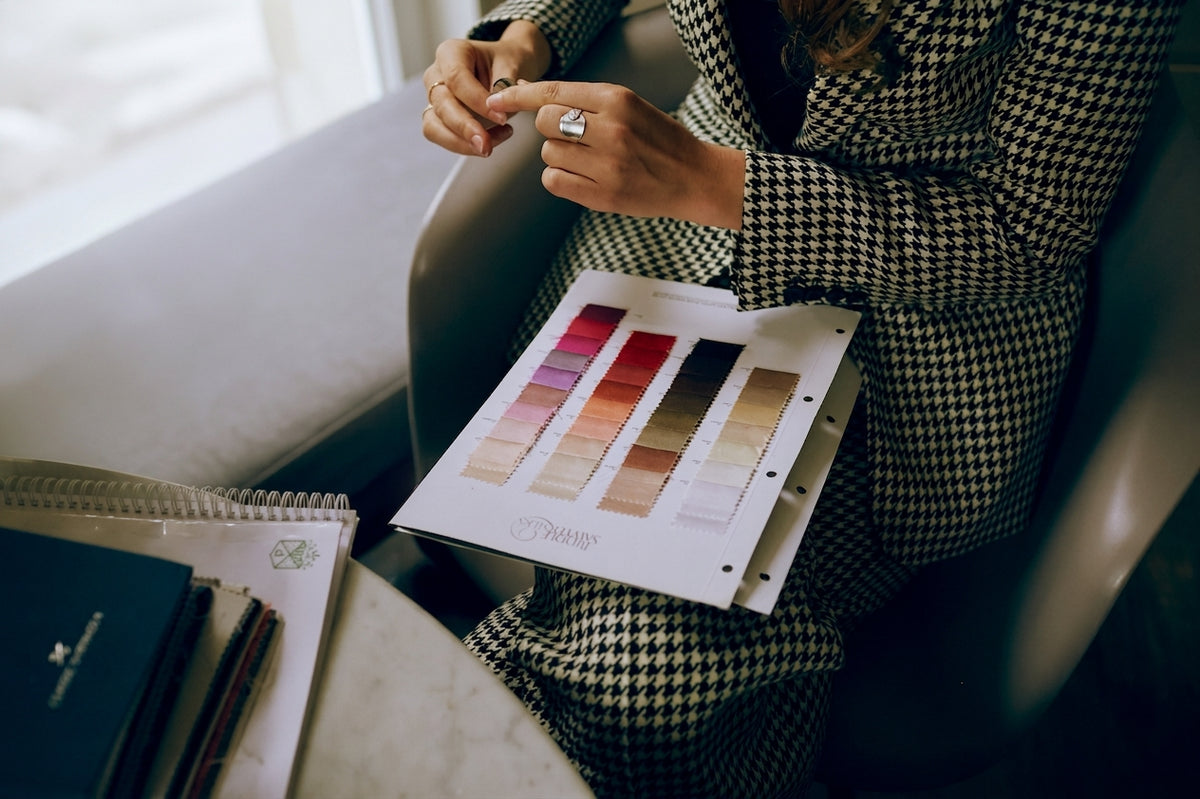 A person in a black-and-white tailored houndstooth suit holds an open fabric colour swatch book showing various colour options, with notebooks on a table nearby.
